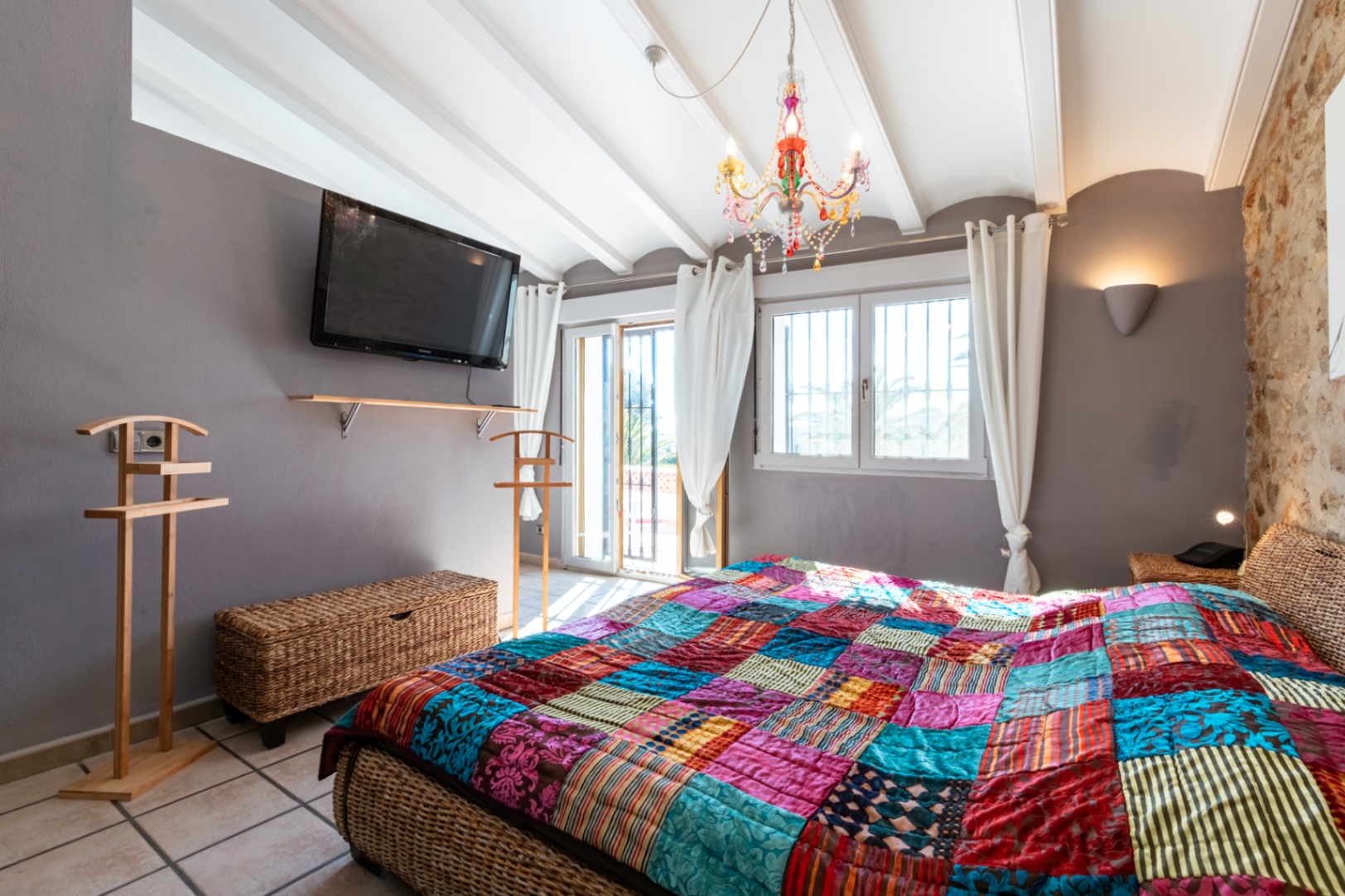 Slaapkamer met natuurstenen muur, wit balkenplafond en een rieten bed. Voorzien van een kleurrijke kroonluchter en directe toegang tot het terras.