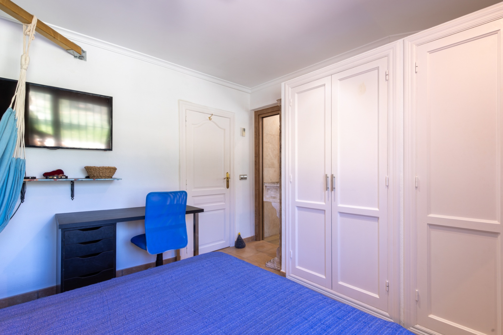 Slaapkamer met witte inbouwkasten, een werkplek met blauwe stoel en toegang tot een aangrenzende badkamer met natuurstenen details.
