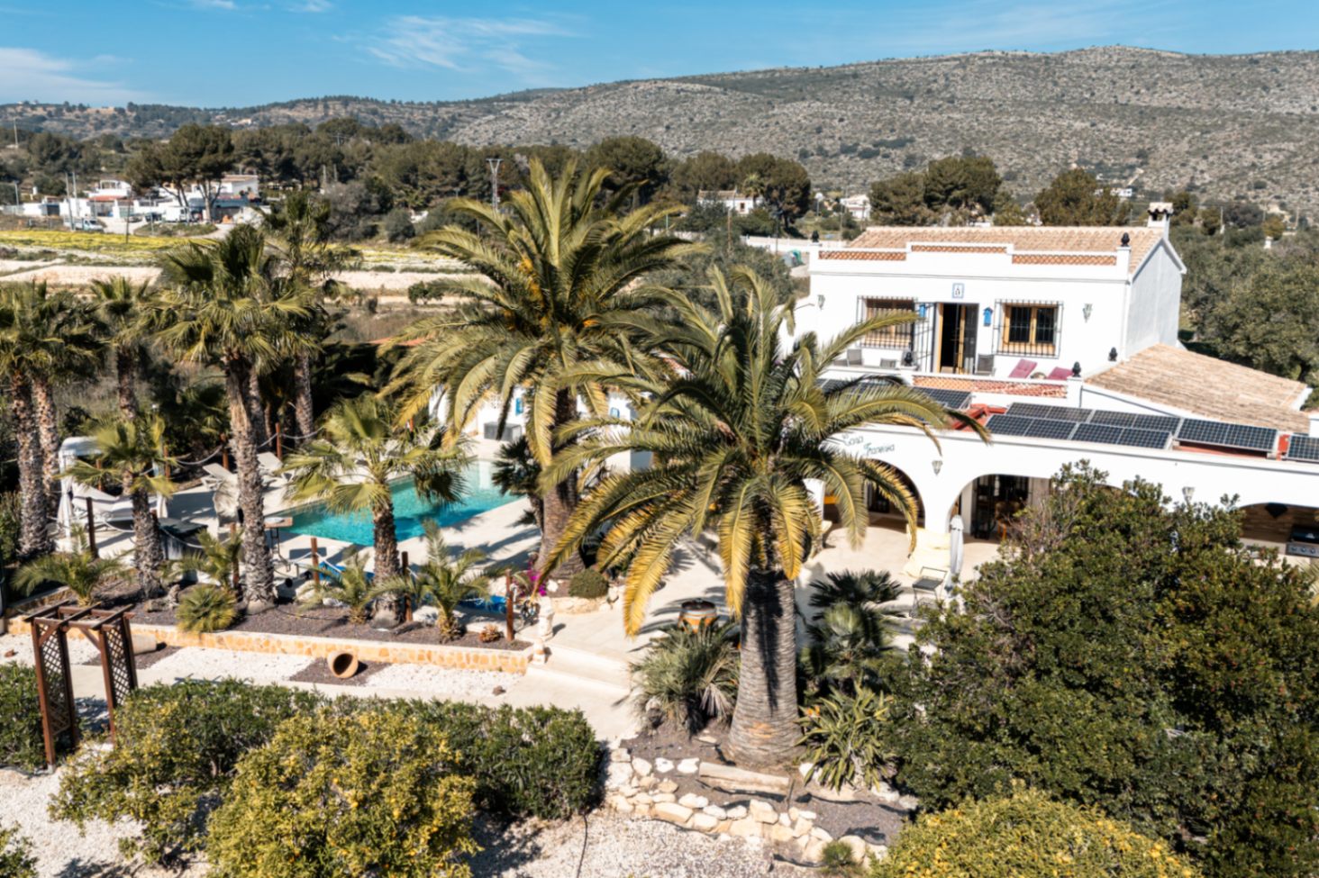 Verhoogd overzicht van de finca met de zonnepanelen, het door palmbomen omgeven zwembad en de traditionele architectuur tegen een bergachtige achtergrond.