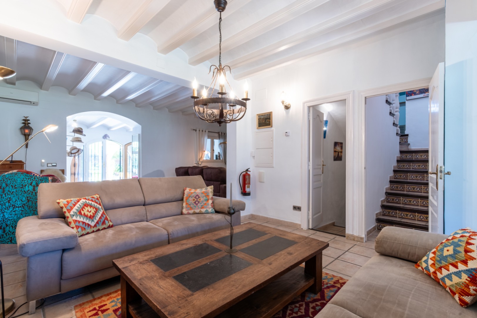 Woonkamer met wit geschilderde balkenplafonds, rustieke tegelvloer en een trap met decoratieve tegels. Voorzien van een massief houten salontafel en smeedijzeren kroonluchter.
