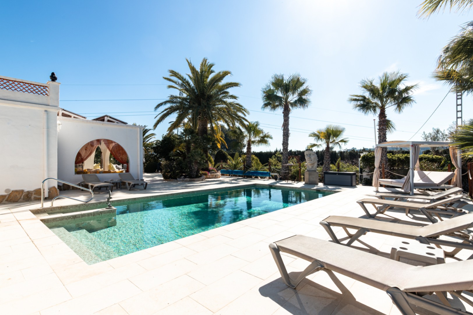 Zwembad van 9,5 x 5 m met een licht stenen terras, ligstoelen, een loungebed en een mediterrane tuin met volwassen palmbomen.