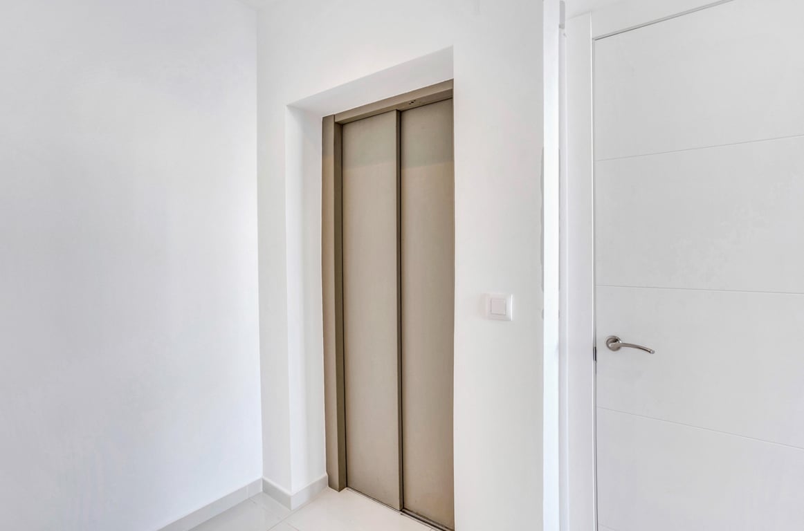 De privé-lift met bronskleurige metalen deuren is geïntegreerd in een minimalistische gang met witte muren en bijpassend binnenschrijnwerk.