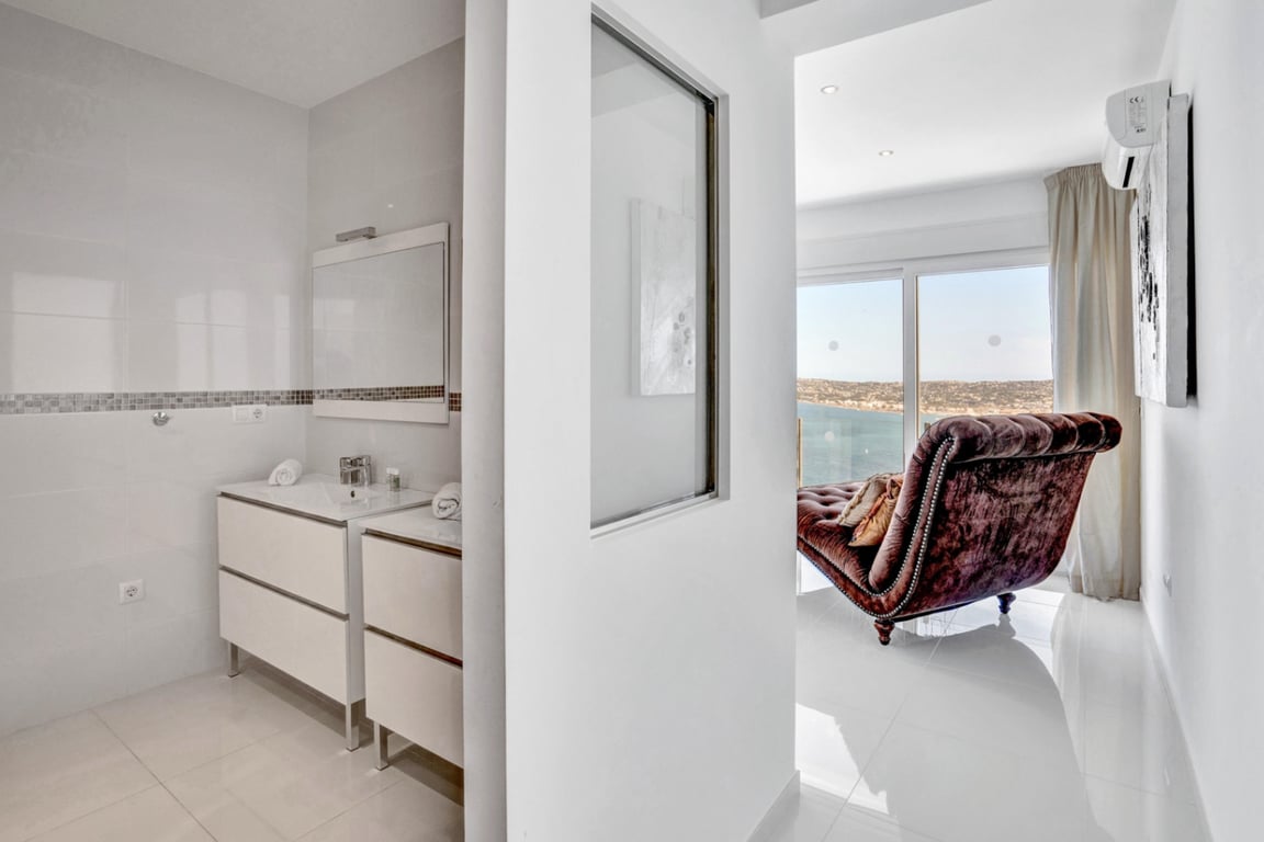 En-suite badkamer met dubbele wastafel en mozaïekaccenten, grenzend aan een lichte kamer met een fluwelen chaise longue en uitzicht op zee.