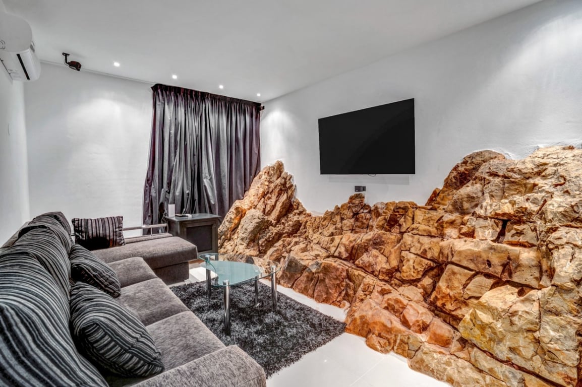 Home cinema kamer met natuurlijke rotswand, grijze bank, glazen salontafel en aan de muur gemonteerde tv met inbouwspots.
