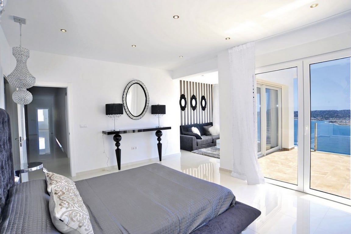 Hoofdslaapkamer met witte gepolijste vloeren, aangrenzende zithoek en glazen schuifdeuren naar het terras met panoramisch uitzicht op zee.