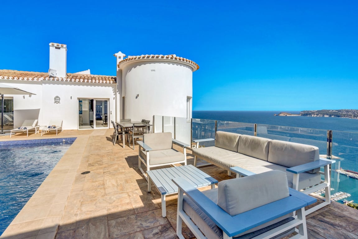 Hoofdterras met loungehoek, eethoek en zwembad, met glazen balustrades die een panoramisch uitzicht bieden op de haven van Jávea en de Middellandse Zee.