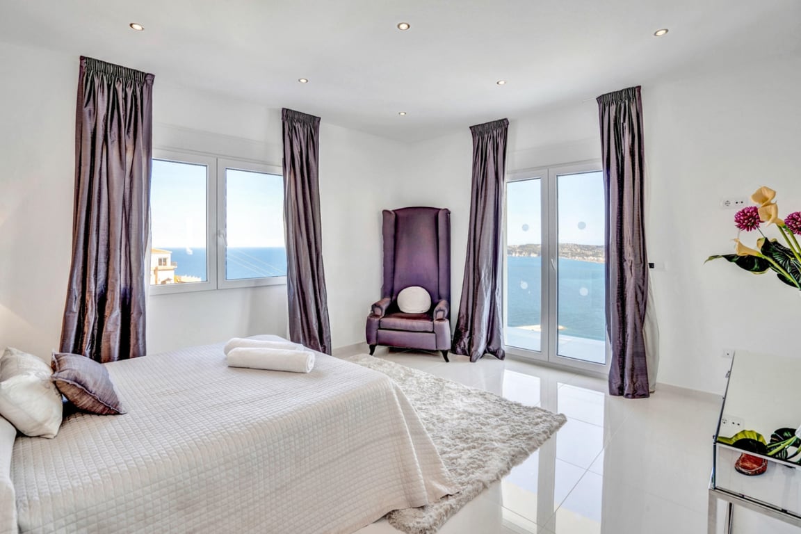Lichte slaapkamer met een paarse fluwelen fauteuil, witte tegelvloer en grote ramen met panoramisch uitzicht over de Middellandse Zee.
