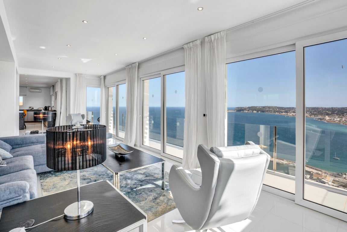 Lichte woonkamer met kamerhoge schuifdeuren die een panoramisch uitzicht bieden op de baai. Voorzien van modern meubilair, witte gordijnen en een strakke tegelvloer.
