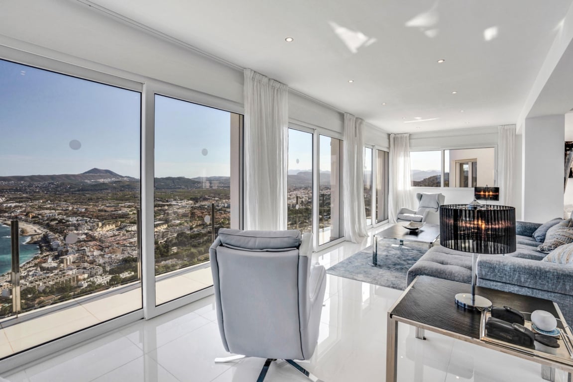 Lichtrijke woonkamer met kamerhoge ramen die een panoramisch uitzicht bieden over de baai van Jávea en de bergen, voorzien van een witte tegelvloer en inbouwspots.