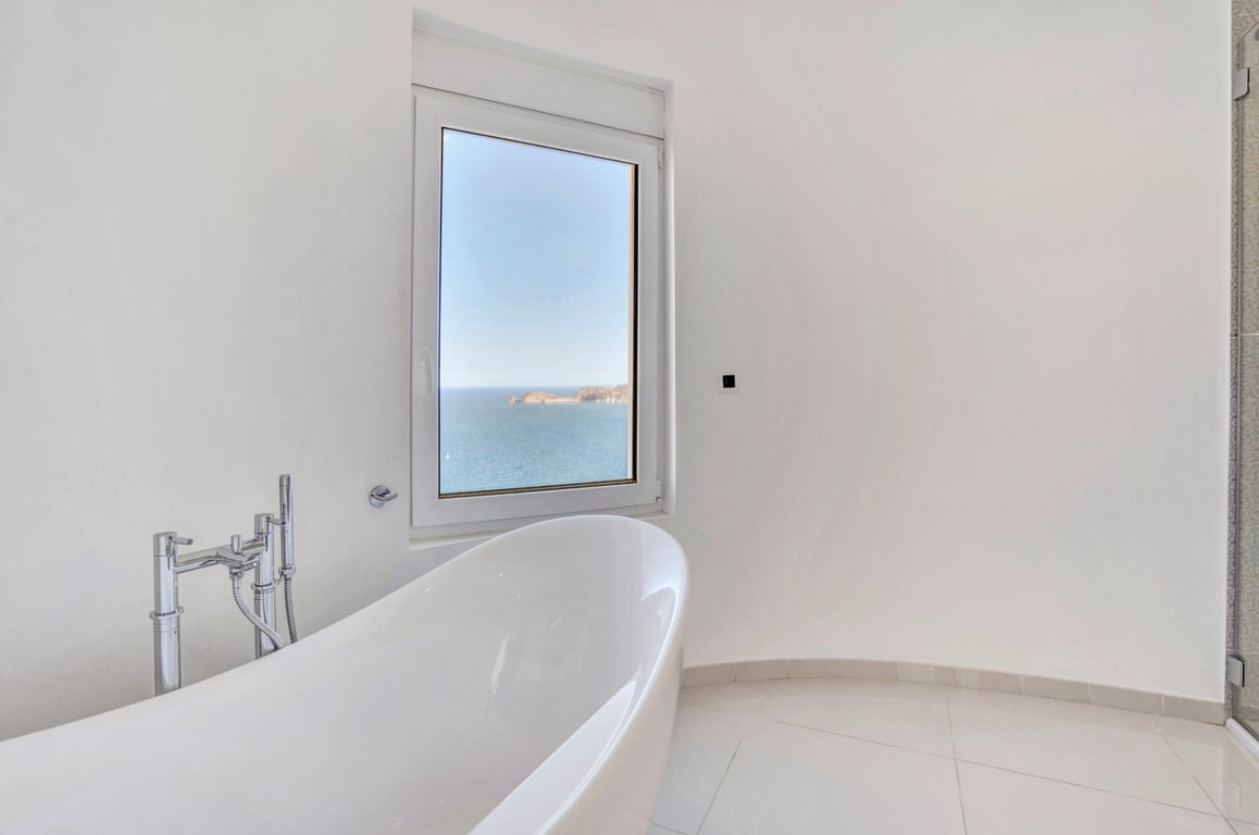Minimalistische badkamer met een vrijstaand wit bad en chromen kranen, met een panoramisch uitzicht op de Middellandse Zee door het raam.