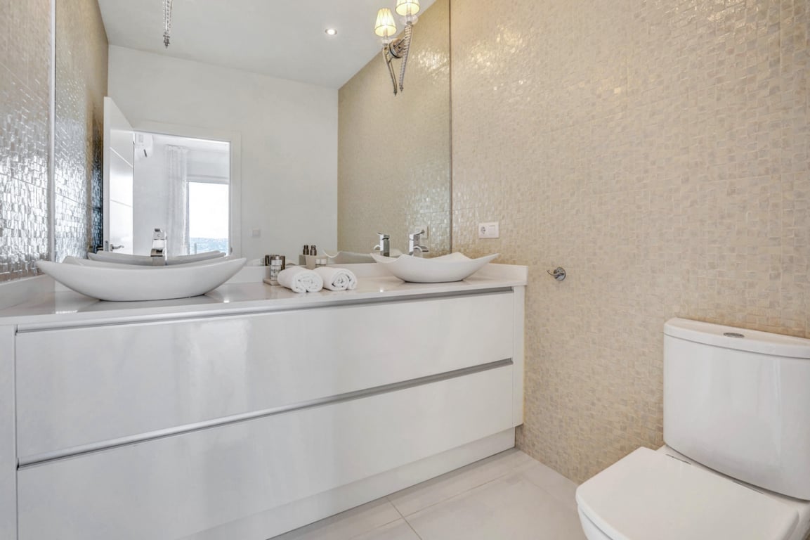 Moderne badkamer met dubbele wastafel op hoogglans wit meubel, beige mozaïekwand en een grote spiegel met wandverlichting.