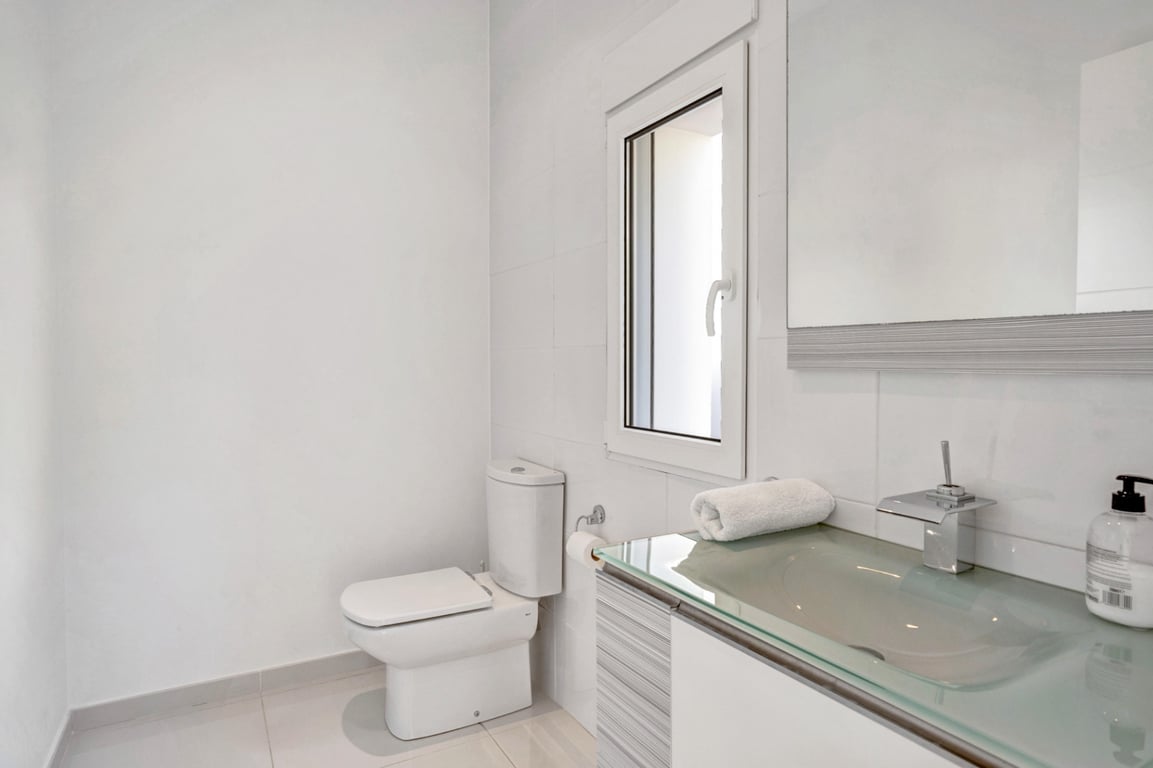 Moderne badkamer met een doorschijnend glazen wastafelblad, chromen kranen en wit betegelde wanden met een klein raam.