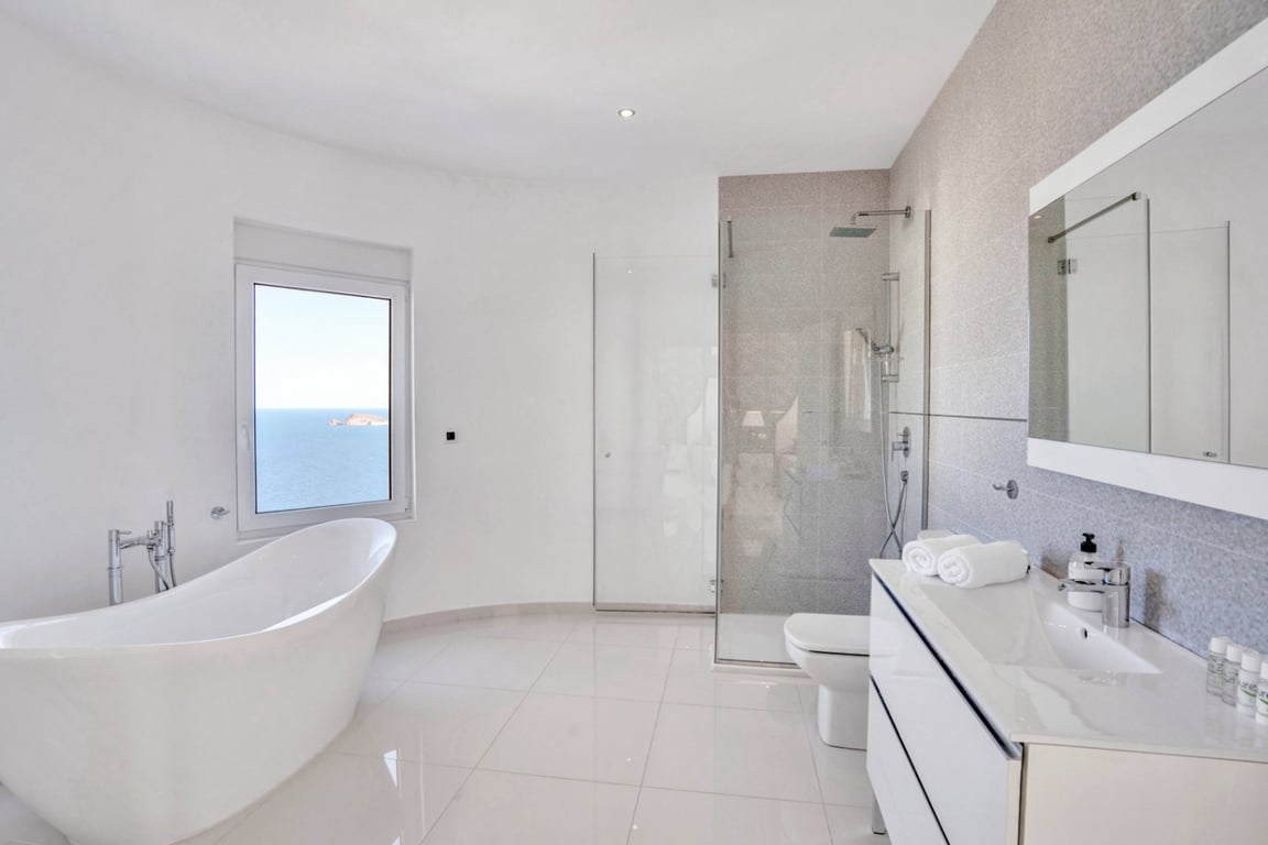 Moderne badkamer met vrijstaand ligbad bij het raam met zeezicht, glazen inloopdouche en wit wastafelmeubel met grote spiegel.