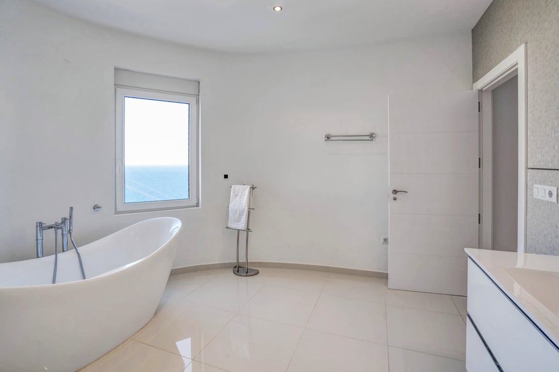 Moderne badkamer met vrijstaand ligbad, lichte tegelvloer en raam met panoramisch uitzicht op de Middellandse Zee.