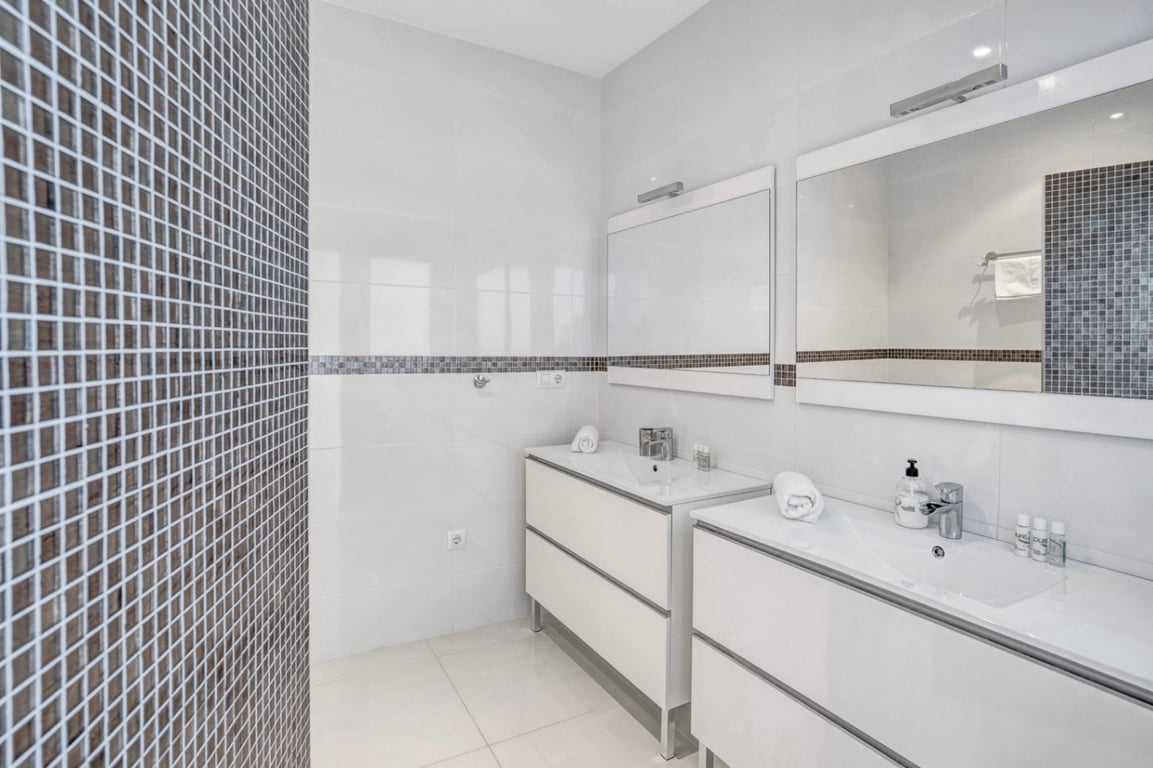 Moderne badkamer voorzien van een dubbele wastafel met wit meubilair, grote spiegels en wanden met donkere mozaïekaccenten.
