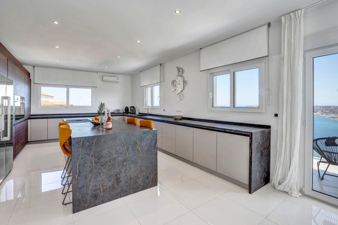 Moderne keuken met een donker natuurstenen eiland, okergele krukken en strakke kasten. De grote raampartijen bieden een panoramisch uitzicht op de Middellandse Zee.