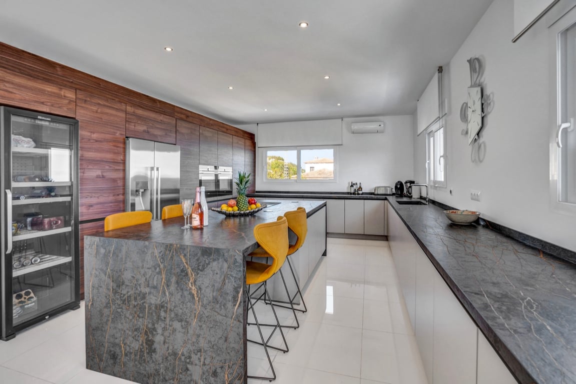 Moderne keuken met een donker stenen kookeiland, gele barkrukken, ingebouwde apparatuur, een wijnkoelkast en houten kastenwanden.