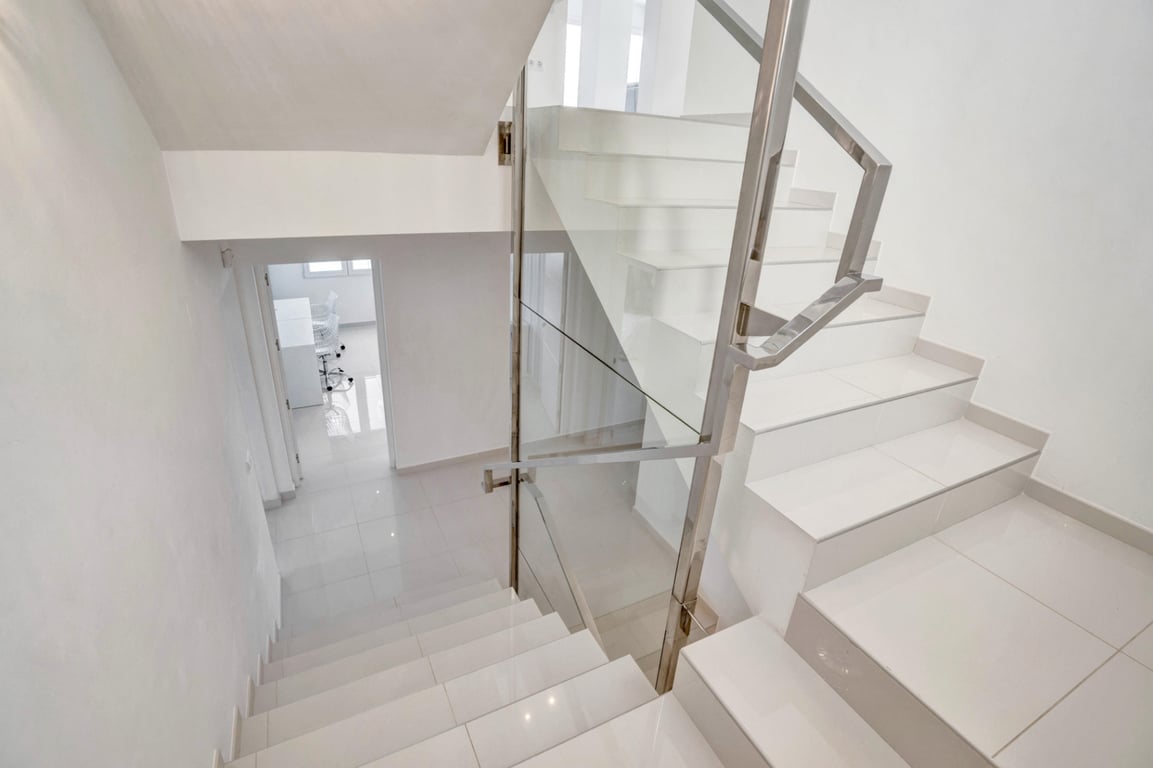Moderne trap met witte betegelde treden, glazen panelen en roestvrijstalen leuningen, passend bij het lichte en minimalistische interieur.