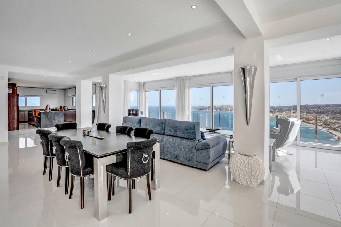 Open woon- en eetkamer met gepolijste vloeren, modern meubilair en grote raampartijen met panoramisch uitzicht op de Middellandse Zee en de baai.