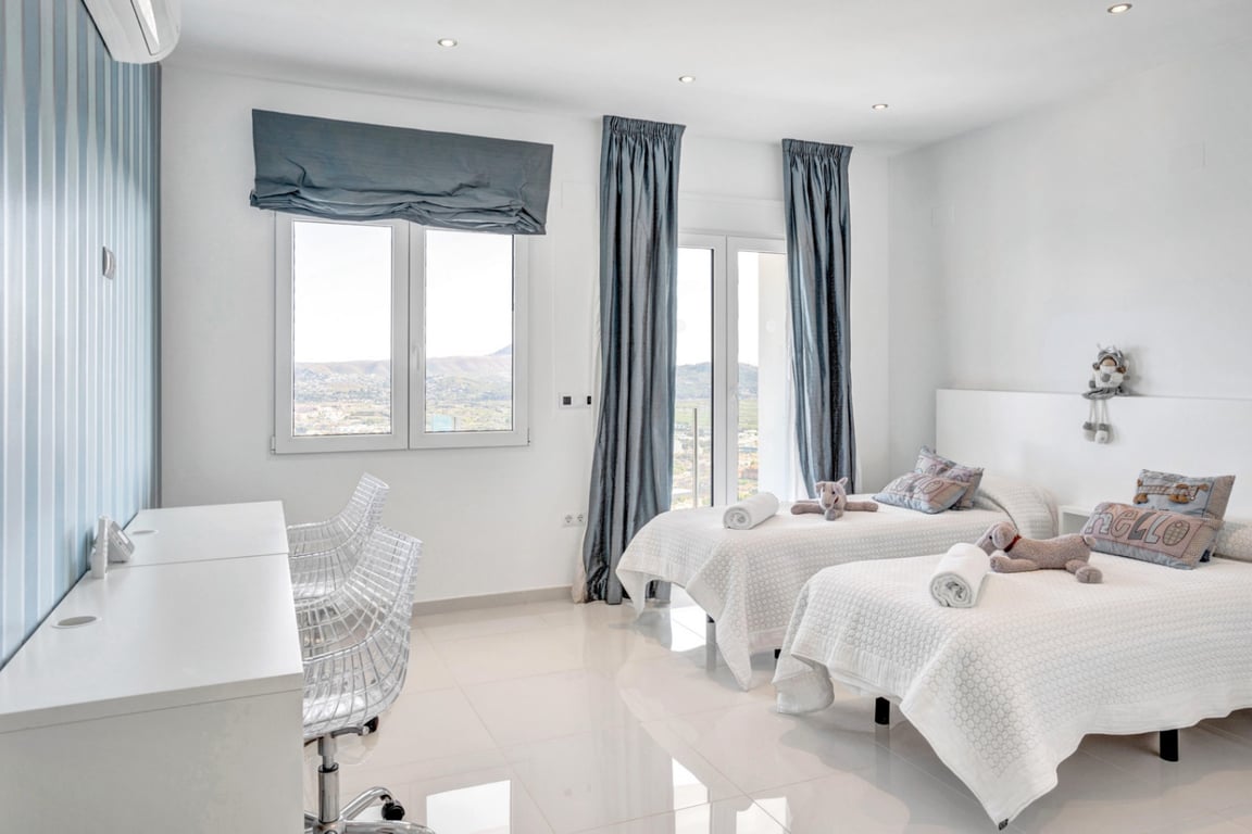 Slaapkamer met twee eenpersoonsbedden, wit bureau met transparante stoelen en grote ramen met panoramisch uitzicht op de bergen en zee.