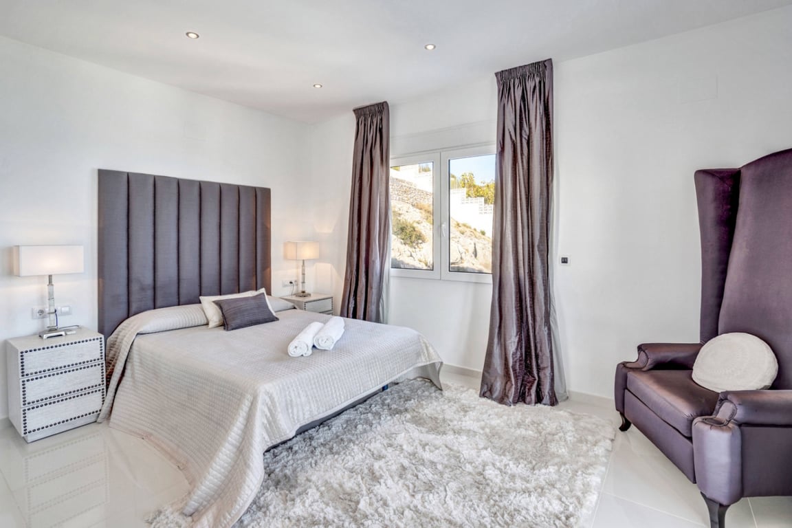 Slaapkamer suite met een hoog grijs hoofdbord, hoogpolig wit tapijt en een paarse fauteuil. Voorzien van inbouwspots en uitzicht op de rotsachtige omgeving.