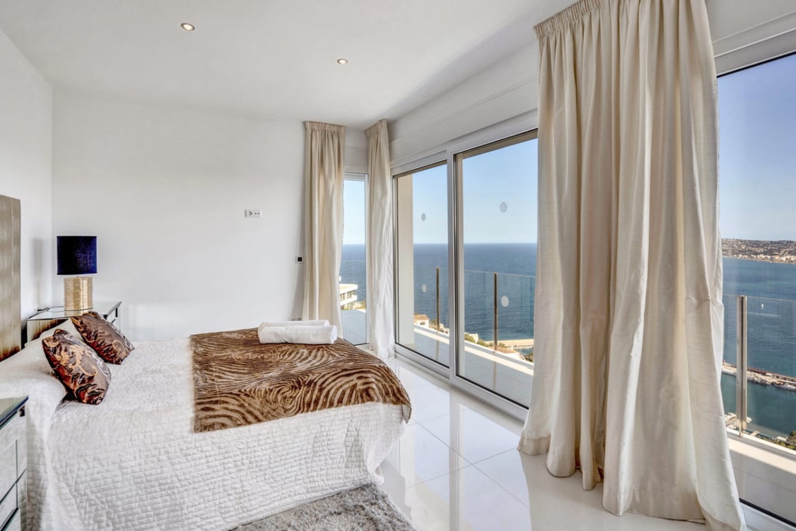 Slaapkamer suite met witte gepolijste tegelvloer, kamerhoge glazen schuifdeuren en direct toegang tot het balkon met panoramisch uitzicht op de Middellandse Zee en de haven van Jávea.