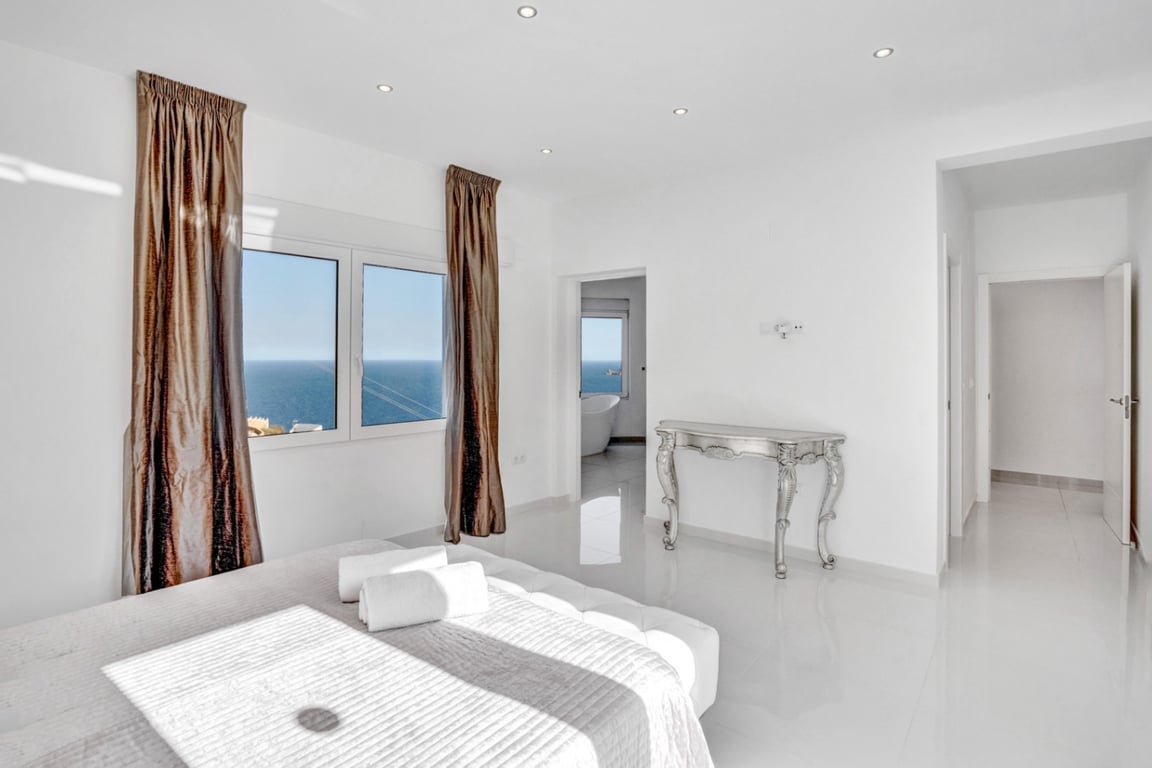 Slaapkamersuite met witte hoogglansvloer, uitzicht op zee en een aangrenzende badkamer voorzien van een vrijstaand ligbad en panoramisch venster.