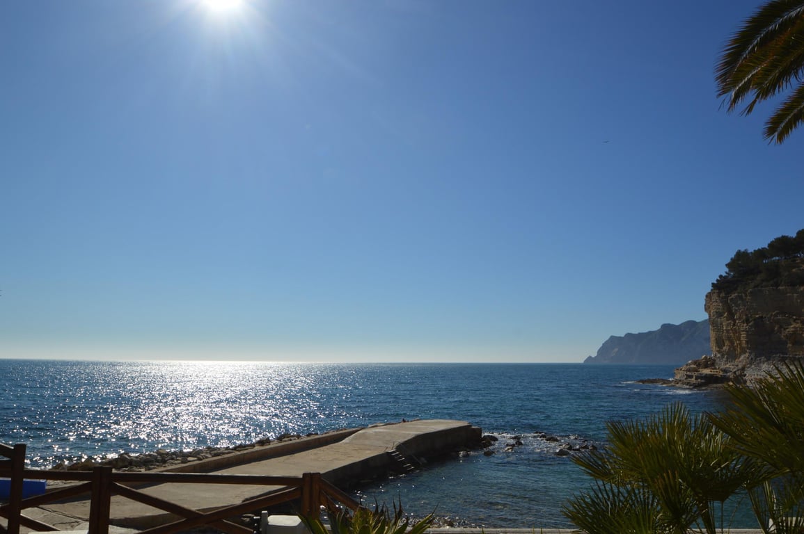 Uitzicht op Cala Advocat met een betonnen pier en glinsterend zeewater. De omgeving wordt gekenmerkt door rotsachtige kliffen en mediterrane palmbomen.