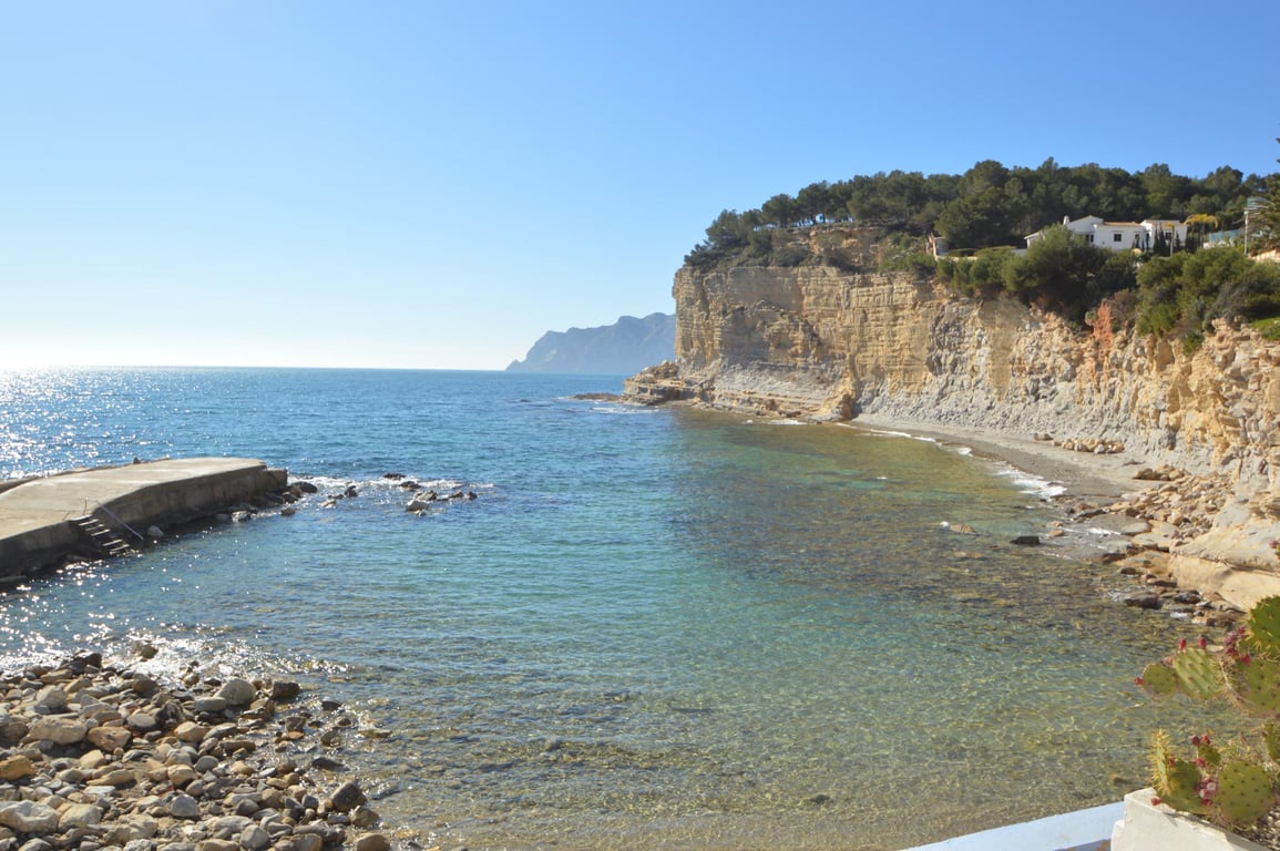 Uitzicht op Cala Advocat met helder turquoise water, een betonnen aanlegsteiger en kalkstenen kliffen begroeid met mediterrane pijnbomen.