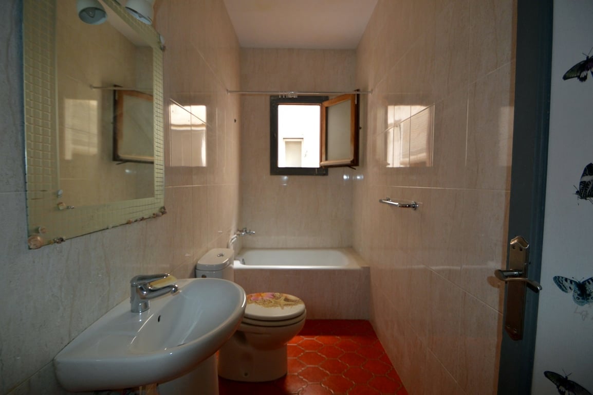 Badkamer met rode zeshoekige vloertegels, beige wandtegels, een wastafel op zuil en een ingebouwd ligbad onder een raam voor natuurlijke ventilatie en lichtinval.