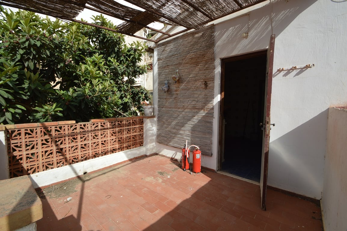 Buitenterras met terracotta tegels, een rieten pergola voor schaduw en een decoratieve muur van keramische windblokken naast volwassen beplanting.