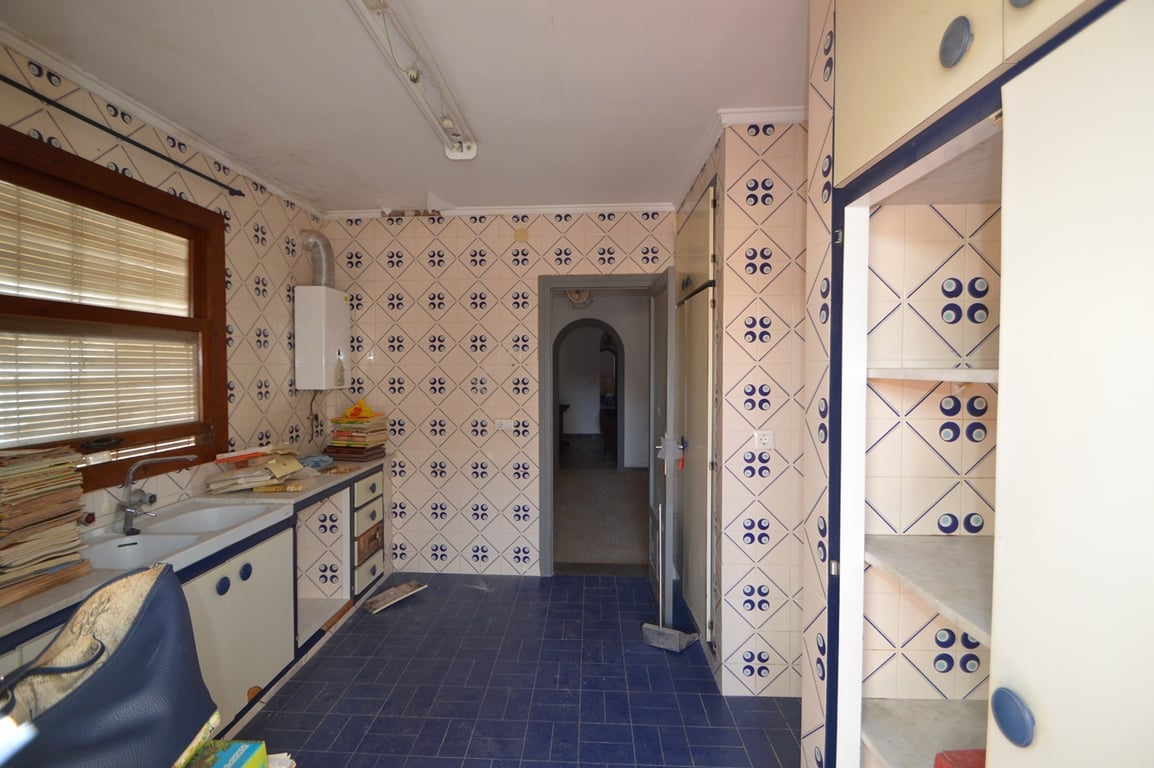 Keuken met gedecoreerde wandtegels, blauwe vloer en witte kasten met blauwe biezen. Voorzien van een dubbele spoelbak onder een houten kozijn en doorgang naar de centrale gang.