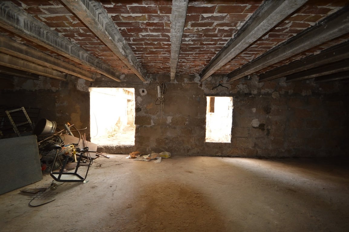 Ruime te renoveren binnenruimte met plafonds van betonnen balken en zichtbare bakstenen. De kamer heeft natuurstenen muren en twee openingen voor natuurlijk licht.