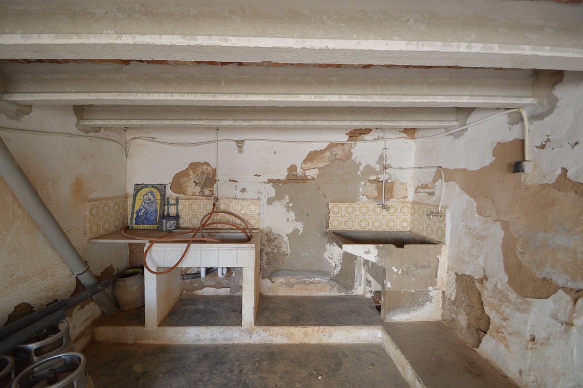 Rustieke bijkeuken met originele stenen spoelbakken, decoratieve tegels en zichtbare balken, perfect voor een volledige renovatie.