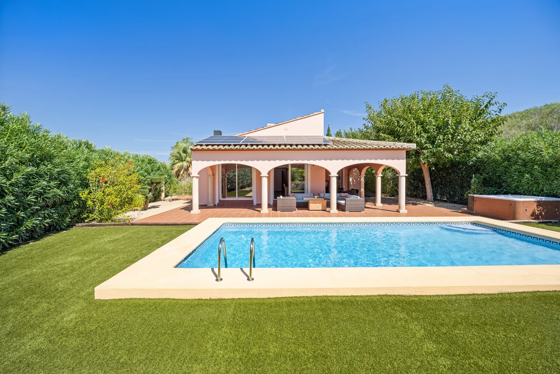 Buitenaanzicht van de villa met mozaïekzwembad, kunstgras en een naya met bogen. Opvallend zijn de zonnepanelen op het dak en de jacuzzi in de tuin.