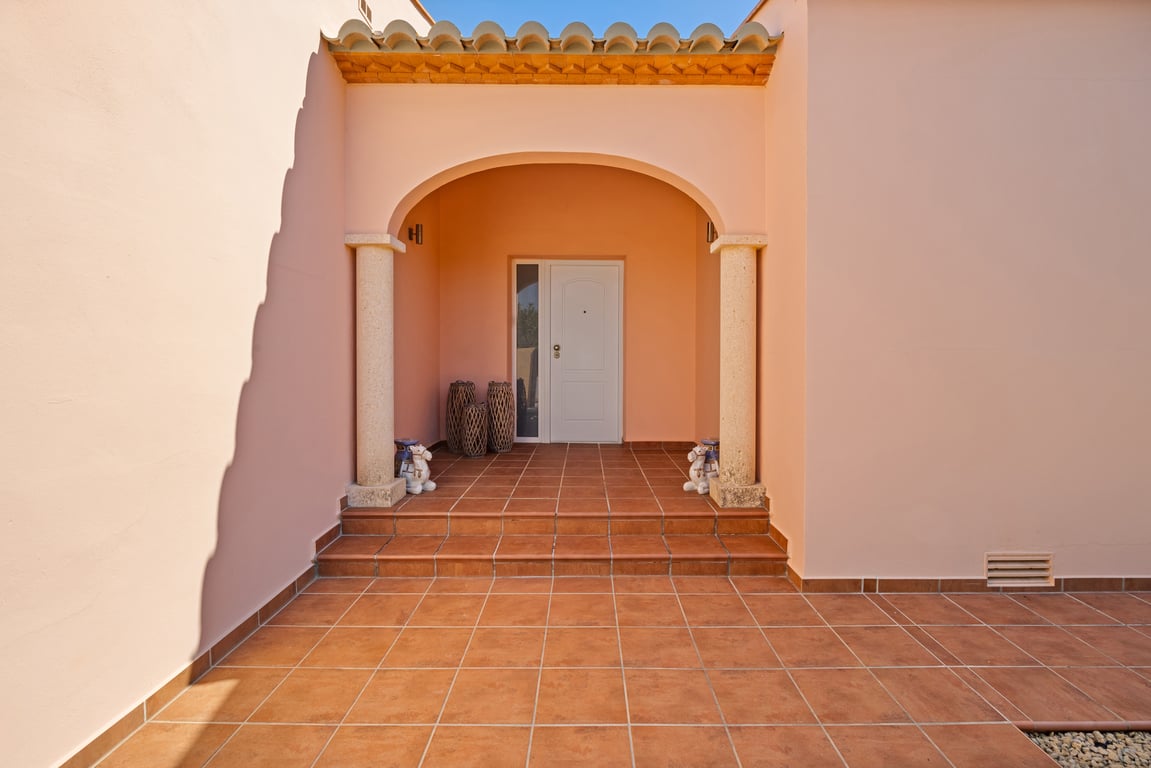 Hoofdingang met een overdekt terras met Toscaanse stenen zuilen en terracotta tegels. De warme gevelkleur en treden leiden naar de witte voordeur met zijraam.