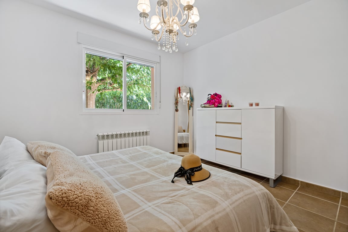 Lichte slaapkamer met tegelvloer, dubbel glas raam met uitzicht op de tuin, centrale verwarmingsradiator en een sierlijke kristallen kroonluchter.