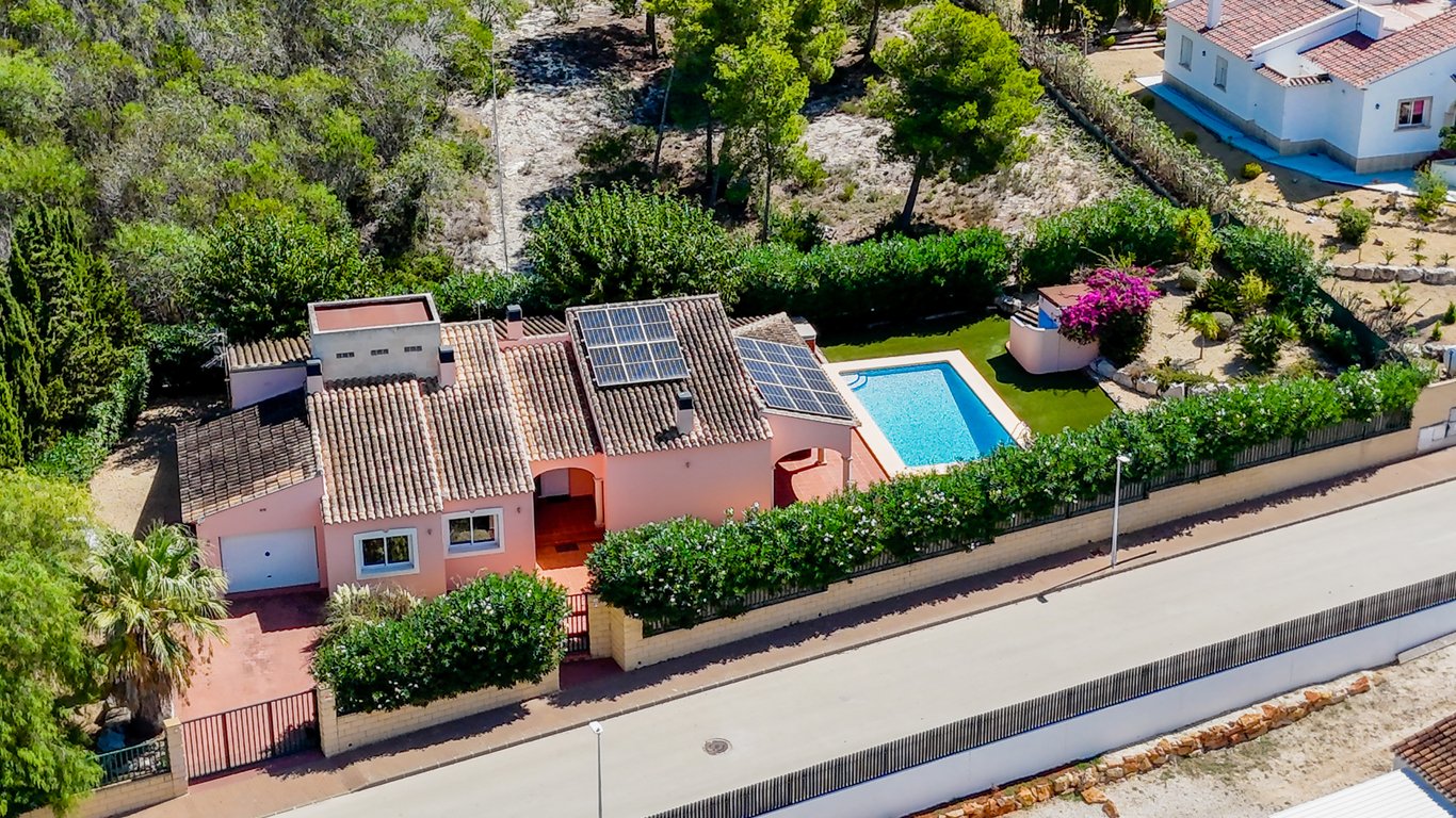 Luchtfoto van de villa met zonnepanelen op het dak, een privézwembad met kunstgras en een volledig omheind perceel met garage en oprit in een rustige omgeving.