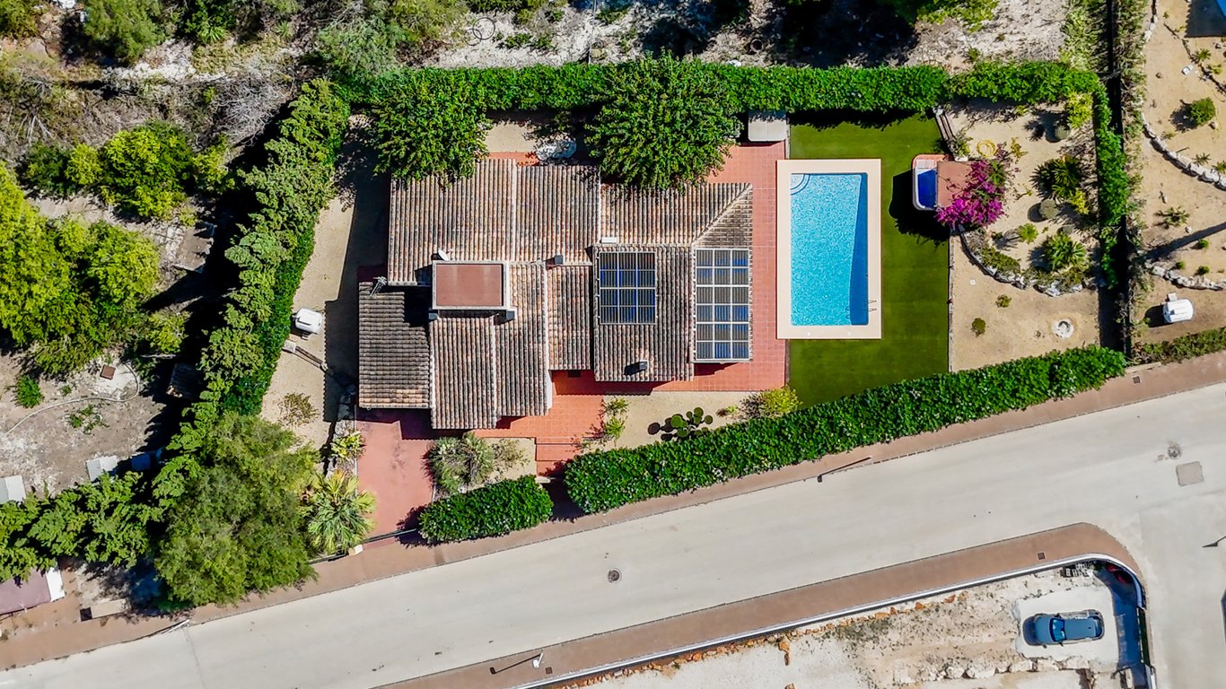 Luchtfoto van de villa met zonnepanelen op het dak, een rechthoekig zwembad, kunstgras en een jacuzzi-gedeelte, volledig omsloten door hoge groene hagen.