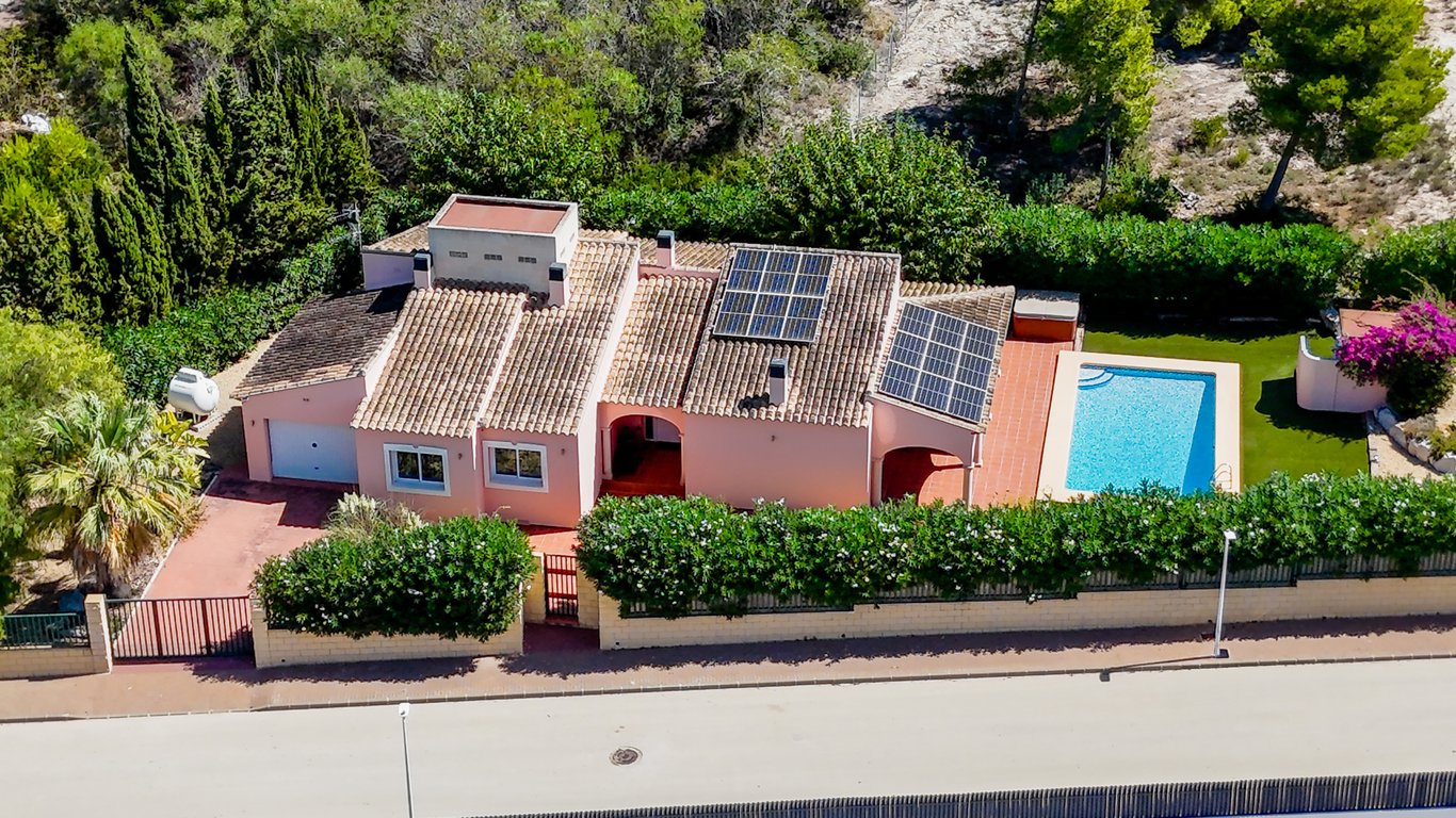 Luchtopname van de gelijkvloerse villa met privézwembad, zonnepanelen op het dak en een volledig omheind perceel met garage en oprit in een groene omgeving.