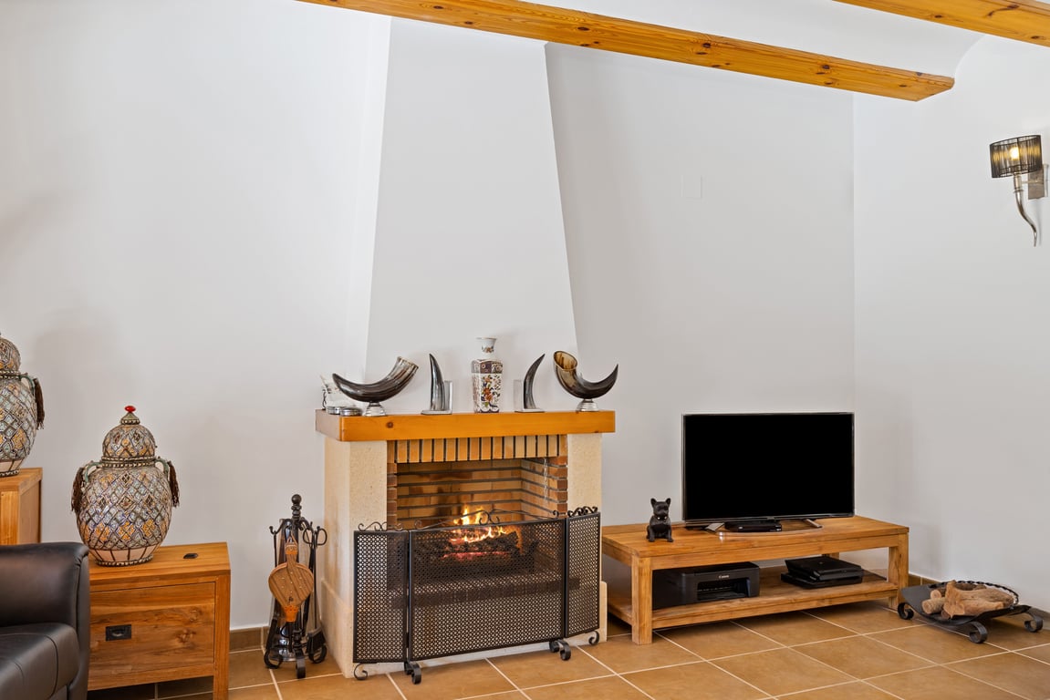 Woonkamer met een traditionele open haard, houten plafondbalken en rustieke vloertegels. Houten meubilair en decoratieve elementen creëren een warme sfeer.