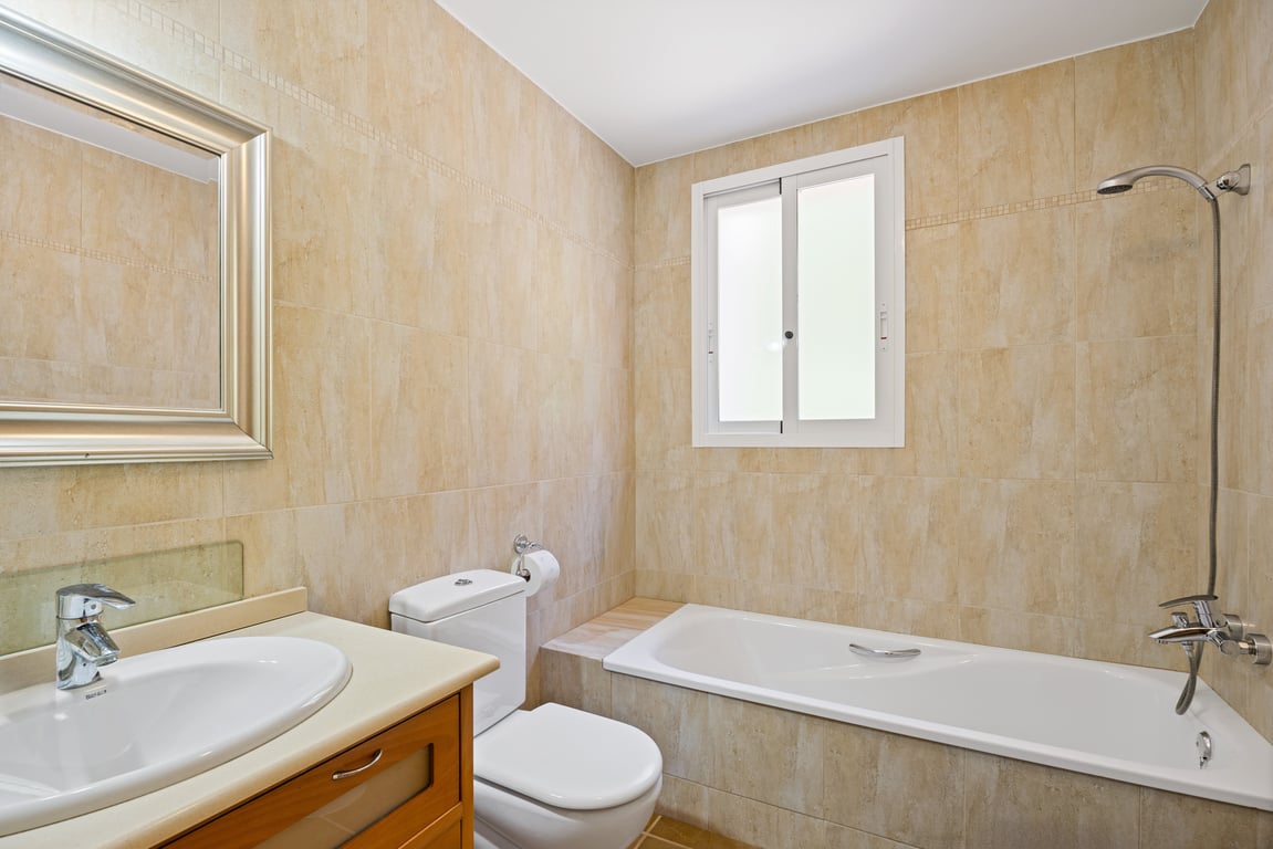 Badkamer met beige betegelde wanden, een ingebouwd ligbad met douchekop, een houten wastafelmeubel en een spiegel met zilveren lijst.