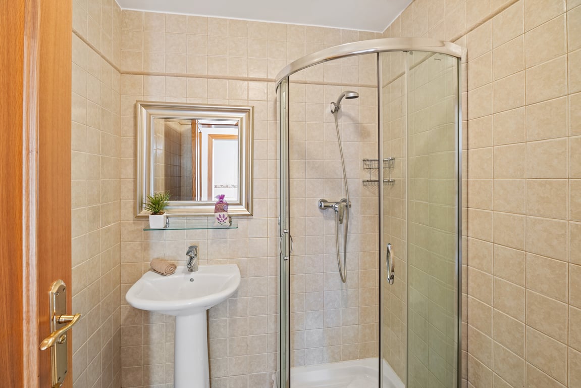 Badkamer met een glazen hoekdouche, een wastafel op zuil en wanden afgewerkt met beige tegels en een subtiele sierrand.