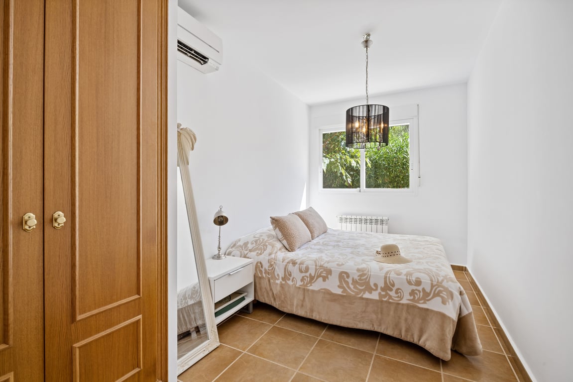 Lichte slaapkamer met een terracotta tegelvloer, split-airconditioning, houten inbouwkast en een raam met uitzicht op het omliggende groen.