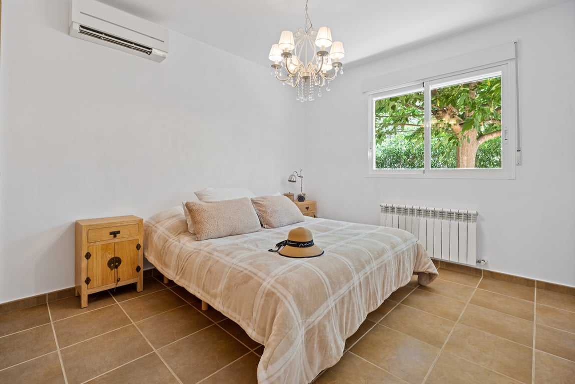 Slaapkamer met tegelvloer, split-airconditioning en radiator. Het raam biedt uitzicht op de tuin, afgewerkt met een klassieke kroonluchter en houten meubels.