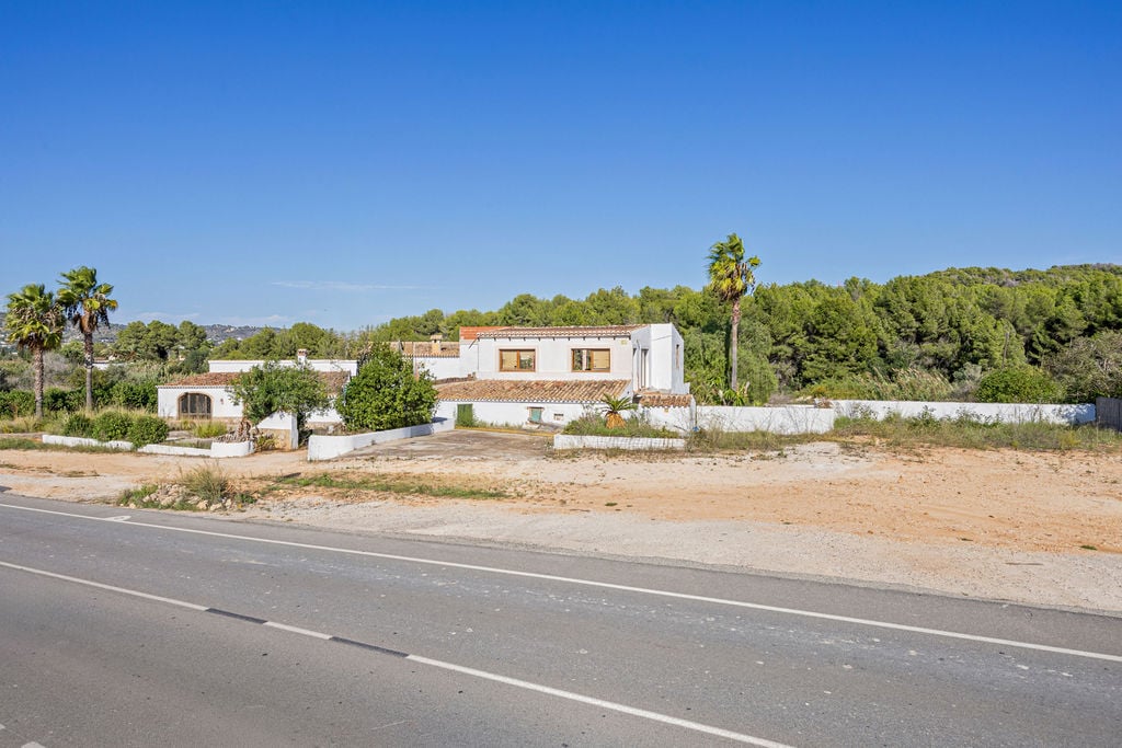 Buitenaanzicht van de traditionele finca met witte gevels en terracotta daken, gelegen aan een toegangsweg en omgeven door mediterrane dennenbossen.