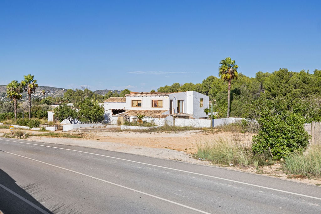 Buitenaanzicht van het pand vanaf de weg, met traditionele witte muren, pannendaken en houten kozijnen in een mediterrane omgeving met pijnbomen en palmen.