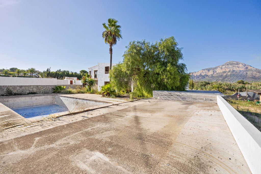 Buitenterras van beton met panoramisch uitzicht op de Montgó, een te renoveren zwembad en een gebouw in mediterrane stijl omgeven door palmen.