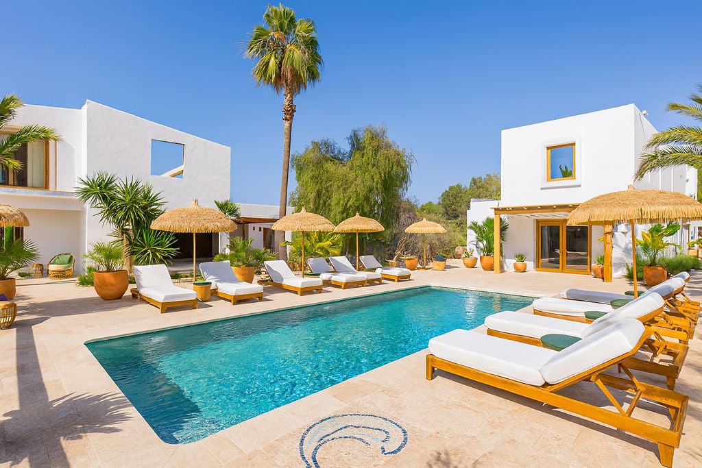 Buitenzwembad met natuurstenen terras, houten ligbedden met witte kussens en rieten parasols, omgeven door witte mediterrane gebouwen.