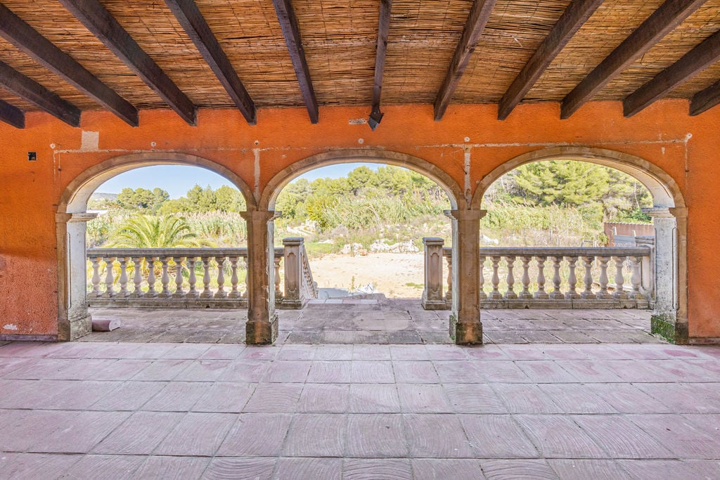 Overdekt terras met stenen bogen, houten balkenplafond met riet en een klassieke balustrade met uitzicht op de omliggende mediterrane natuur.